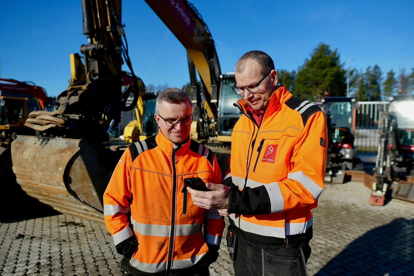 To anleggsarbeidere ser på mobilen foran maskinparken